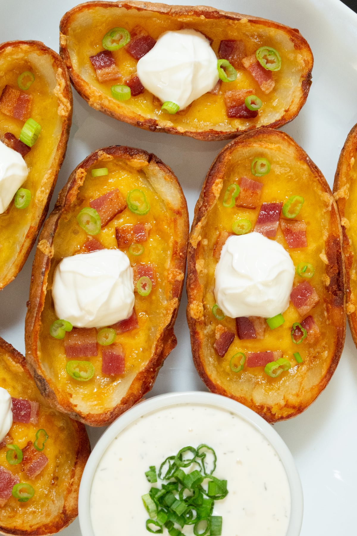 Several potato skins on a white serving dish next a full ramekin of ranch dressing that's topped with green onions.